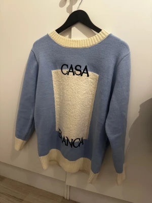 Blå stickad tröja Casablanca M - Snygg ljusblå stickad tröja från Casablanca med kontrasterande vita ribbade muddar och krage. Framsidan har ett stort vitt fluffigt parti med broderad text 'CASA BLANCA'. Perfekt för dig som gillar unik design och mjuka material.