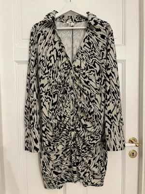 Zebramönstrad kortklänning L - Snygg kortklänning med zebramönster i svart och beige. Klänningen har lång ärm, krage och knappar framtill med rynkade detaljer längs knappraden. Materialet är mjukt och följsamt, perfekt för dig som gillar statement-plagg med coola mönster.