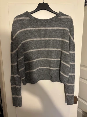 Gråvit randig stickad tröja Divided - Mysig grå stickad tröja från Divided med vita ränder. Tröjan har en avslappnad passform, ribbad rund hals och långa ärmar. Perfekt för kyliga dagar.