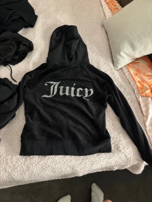 Svart Juicy Couture velour hoodie - Svart hoodie från Juicy Couture i mjuk velour med glittrande stenar som bildar 'Juicy' på ryggen och ett J på bröstet. Jackan har huva, snörning, framfickor och dragkedja med metall-J. Perfekt för dig som gillar klassisk Y2K-stil. Pris går att diskutera.🥰