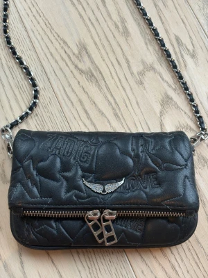 Svart axelväska från Zadig & Voltaire - Cool svart axelväska i läder från Zadig & Voltaire med quiltat mönster av hjärtan, stjärnor och blixtar. Väskan har silverfärgade detaljer, kedjerem med insytt läder och dubbla dragkedjor framtill. Snygg vingedetalj med stenar på framsidan.