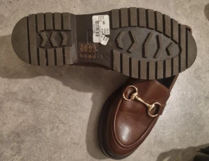 Bruna loafers med gulddetalj från XIT - Snygga bruna loafers från XIT med chunky svart sula och guldfärgad metallspänne över foten. Skorna har rund tå och är tillverkade i skinnimitation med en klassisk, stilren look. Perfekta för dig som gillar tidlös design med en modern twist.