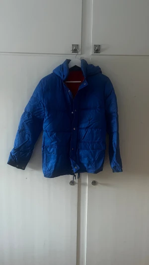Blå pufferjacka med huva - Säljer en äldre blå pufferjacka med huva och dragkedja samt knappar framtill. Jackan har två fickor och justerbar nederkant med snörning. Perfekt för kalla dagar och har en schysst loose passform som ger en chill vibe.