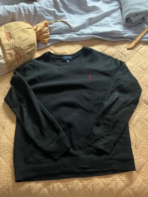 Svart sweatshirt från Polo Ralph Lauren - Svart sweatshirt från Polo Ralph Lauren med klassisk rund hals och långärmad design. Tröjan har en liten röd Polo-logga broderad på bröstet. Tillverkad i mjuk bomull och polyesterblandning, perfekt för chill dagar. Enkel och stilren look som funkar till jeans eller joggers.