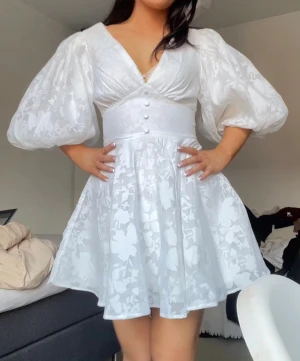 Vit kortklänning med puffärmar från Showpo - Superfin vit kortklänning med v-ringning, markerad midja och dekorativa knappar framtill. Klänningen har stora puffärmar och ett subtilt blommigt mönster i tyget. Kjolen är utsvängd och materialet känns lätt och luftigt, perfekt för sommarens alla tillfällen. Endast använd en gång. Perfekt för student! Nypris 1200