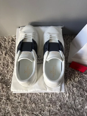 Valentino Garavani vita sneakers - Snygga vita Valentino Garavani sneakers med svart detalj över snörningen. Klassisk rund tå, platt sula och diskret logga på insidan. Tillverkade i skinn och har en stilren, lyxig vibe. Perfekta för dig som vill ha något exklusivt men ändå cleant.