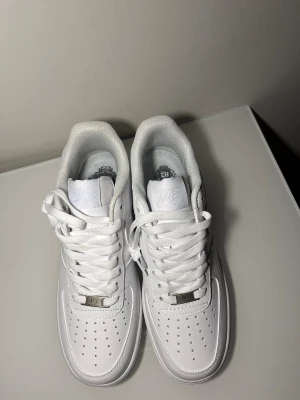 Nike Air Force 1 helvita sneakers - Klassiska Nike Air Force 1 i helvitt skinn med vit sula och snörning. Ikonisk siluett med perforerad tå och broderad Nike Air-logga på hälen. Helt nya och oanvända – säljes eftersom de tyvärr blev för små.