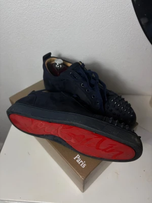 Christian Louboutin blå mocka med slickar sneakers - Säljer ett par mörkblå sneakers från Christian Louboutin i mocka med ikoniska röda sulor och coola nitar på tån.(saknar ett par sprinar syns på bilden) Skorna har rund tå, snörning och en platt sula. Perfekt för dig som vill sticka ut med en lyxig och edgy stil.