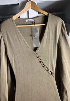 Beige långklänning från Adoore - Långklänning i beige från Adoore med v-ringning och omlott-detalj framtill, dekorerad med små knappar. Klänningen har långa puffärmar med resår vid handlederna och är tillverkad i ett mjukt, stretchigt material som sitter snyggt på kroppen.