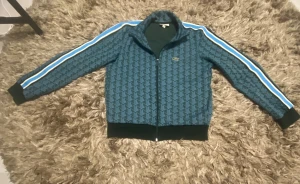Grön mönstrad track jacket från Lacoste - Snygg grön track jacket från Lacoste med geometriskt mönster och klassisk krokodillogga på bröstet. Jackan har svarta muddar, ståkrage och dragkedja framtill. Längs ärmarna går vita och blå ränder som ger en sportig vibe. Perfekt för dig som gillar streetwear.