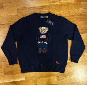 Mörkblå stickad tröja Polo Bear - Mörkblå stickad tröja från Polo Ralph Lauren med ikonisk Polo Bear broderad på bröstet och RL-broderi i rött vid nederkanten. Rund hals och ribbade muddar. Tröjan är gjord i mjuk bomull och har lång ärm, perfekt för dig som vill ha en klassisk men lekfull look.