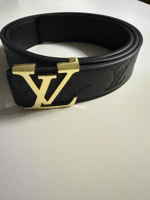 Svart Louis Vuitton skärp med guldspänne - Louis Vuitton bälte i svart skinn med präglade LV-loggor och monogramdetaljer längs hela remmen. Snyggt och brett bälte med ett rejält guldspänne i metall med LV-logotypen framtill. Perfekt för dig som vill ha en lyxig och stilren accessoar.