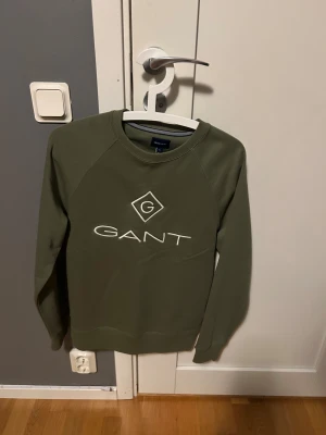 Olivgrön sweatshirt från GANT - Snygg olivgrön sweatshirt från GANT med broderad logga i vitt framtill. Tröjan är använd en gång och är i nytt skick Storlek S. Hör av dig vid minsta fundering!