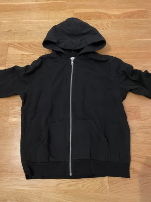Svart hoodie med dragkedja - Svart hoodie med hel dragkedja framtill och två stora fickor. Tröjan har huva och är långärmad, perfekt för chill dagar. Materialet känns mjukt och skönt, troligen bomull eller polyester. Enkel och stilren design utan mönster eller logga.