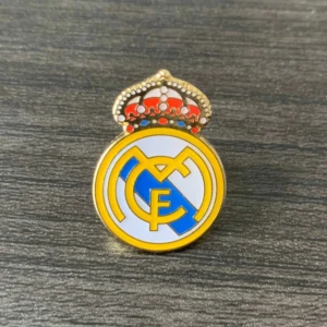 Real Madrid pin i metall - Snygg brosch med Real Madrid-loggan i guld, vitt, blått och rött. Broschen har en rund form med en krona upptill och detaljerade färger som gör den riktigt iögonfallande. Perfekt för dig som vill visa ditt stöd för klubben. Den är i metall och passar att sätta på jacka, väska, kavaj eller keps! 