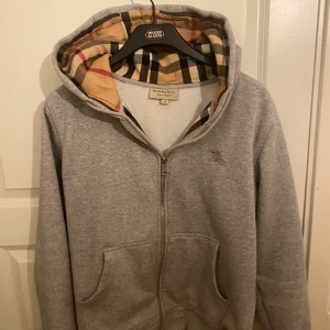 Grå Burberry hoodie med rutig huva - Säljer en grå hoodie från Burberry i storlek M med klassisk rutig insida på huvan. Tröjan har dragkedja, snörning vid huvan och broderad Burberry-logga på bröstet. Mjuk bomullskänsla och två fickor framtill. Perfekt för dig som gillar stilrena detaljer. Tröjan är i bra skick. Om du är intresserad för fler bilder kom gärna privat. Öppen för prisförslag.