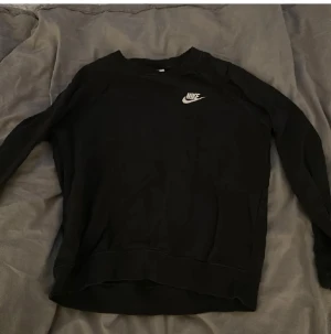 Svart Nike sweatshirt med logga - Svart sweatshirt från Nike med klassisk vit logga på bröstet. Tröjan har rund hals och långa ärmar, perfekt för chill dagar eller när du vill ha en enkel och snygg look. Mjuk bomullskänsla och normal passform.