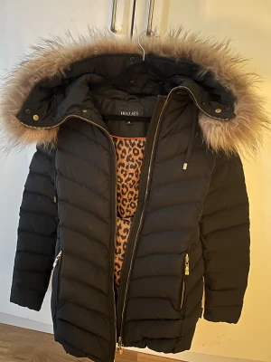 Svart pufferjacka Hollies med päls - Svart pufferjacka från Hollies med fluffig päls på huvan och leopardmönstrat foder. Jackan har guldfärgade dragkedjor, långa ärmar och snygga detaljer som ger en cool vibe. Perfekt för kalla dagar när du vill vara både varm och stylish. obs skärp på jackan saknas men den är lika fin.