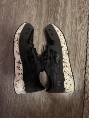 Svarta Asics sneakers med mönstrad sula - Svarta sneakers från Asics med coola detaljer och snörning. Skorna har en vit sula med svarta och orangea stänk som ger en unik look. Materialet är syntet och mesh för bra andning och komfort. Perfekta för dig som gillar sportig stil.