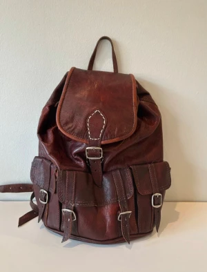 Läderryggsäck / Skórzany plecak - Ryggsäck i mörkbrunt läder med flera fickor och justerbara remmar. Oanvänd, i nyskick med silvriga detaljer. Bredd 28 cm Höjd 36 Banden / remmarna är 78 cm långa Kategori Varumärke Skick Färg Uppladdad Ryggsäckar