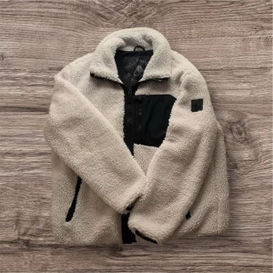 Moose Knuckles SAGLEK ZIP - Säljer en vit teddyjacka från Moose Knuckles med svart bröstficka och detaljer. Jackan har hög krage, hel dragkedja och två sidofickor med svarta dragkedjor. Materialet är mjuk polyester med bomullsfoder, perfekt för kyliga dagar. Inga övriga defekter förutom den som vissas på sistas bilden. Nypris ligger upp mot 5500 säljer den nu för ett bra pris, håller värme i denna kyla, kom privat för mer frågor!