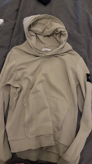 Beige hoodie från Calvin Klein Jeans - Snygg beige hoodie från Calvin Klein Jeans med stor huva och ribbade muddar. Tröjan har en svart CK-logga på vänster ärm och en praktisk magficka framtill. Perfekt för dig som gillar stilrena och bekväma plagg.skeiv gärna vid frågor.pris kan diskuteras.