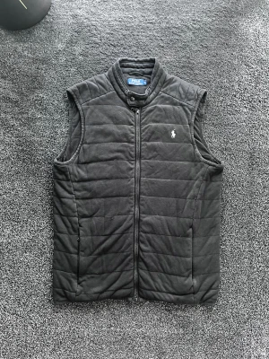 Ralph Lauren väst - Svart quiltad dunväst från Polo Ralph Lauren med ståkrage och dragkedja framtill. Västen har ett broderat vitt Polo-logotyp på bröstet och en knappdetalj vid halsen. Perfekt att slänga över en hoodie för en chill look. Skick 8/10, storlek M. Pris går att diskutera✅