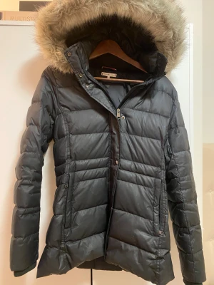 Svart dunjacka med päls från Tommy Hilfiger - Svart dunjacka från Tommy Hilfiger med fluffig päls på huvan och snygga quiltade detaljer. Jackan har dragkedja, knappar och två fickor framtill. Insidan har Hilfigers klassiska ränder i rött, vitt och blått. Perfekt för kalla dagar. (Köptes som present och har bara använts 1 gång!) pris kan diskuteras. 