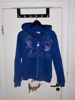 Blå hoodie Aim For The Moon S - Blå hoodie med glittriga paljettdetaljer och tryck framtill, dragkedja och huva. Mjuk insida i fleece och praktiska fickor. Snyggt motiv med text och figur på bröstet, perfekt för dig som gillar statement-plagg.