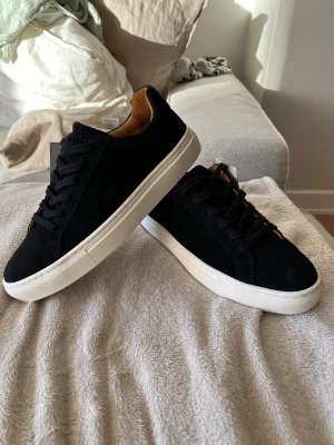 Svarta mockasneakers från A Day's March - strl 42 - Snygga svarta sneakers i mocka från A Day's March med klassisk låg profil och vit platt sula. Skorna har svarta snören och en stilren design med brunt innerfoder. Perfekta för dig som gillar minimalistisk och clean stil. Köpta på deras sample sale av flickvännen men var tyvärr för små för mig. Ordinare pris på deras hemsida: 1795 SEK