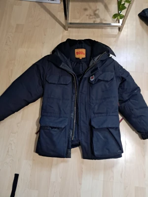 Svart vinterjacka från Fjällräven XXXS - Säljer en svart vinterjacka från Fjällräven i storlek XXXS. Jackan har flera stora fickor med lock, dragkedja och snörning vid huvan. Insidan är quiltad och jackan har klassisk Fjällräven-logga i nacken. Perfekt för kalla dagar och outdoor vibes. Skulle säga är en normal S liten M. Snörenavid huvan har elasticen försämrats se bild