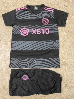 Inter Miami Messi fotbollströja set - Svart och grå fotbollströja med rosa detaljer från Inter Miami, Messi #10 på ryggen. Kortärmad med unikt mönster och klubbmärke på bröstet. Matchande shorts med klubbmärke. Tillverkad i lätt och ventilerande material, perfekt för fotbollsträning.