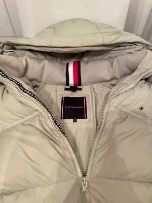 Säljer nu min ljusgröna Tommy hilfiger! Riktig bra jacka under vintern  - Snygg beige pufferjacka från Tommy Hilfiger med huva och broderad logga på ryggen. Jackan har dragkedja framtill, två fickor och klassisk Hilfiger-detalj på ärmen. Perfekt för kalla dagar och har en stilren look med quiltad design.