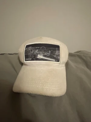 Vit keps från OneCap - Säljer en beige keps från Onetap med ett svartvitt stadsmotiv framtill. Kepsen har en mjuk, nästan plyschig yta och böjd skärm. Perfekt accessoar för dig som vill sticka ut med en unik och stilren look.