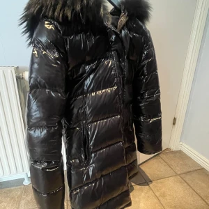 Svart glansig dunjacka med pälskrage - Säljer en svart, glansig dunjacka med stor pälskrage och dragkedja framtill. Jackan är lång och har fickor med dragkedja. Perfekt vår jacka. 