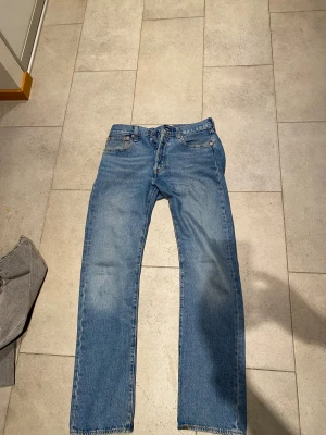 Levi's 501 blå jeans W32 L34 - Klassiska Levi's 501 jeans i ljusblå denim med raka ben och femficksmodell. Byxorna har knappgylf och den ikoniska läderpatchen bak i midjan. Perfekta för dig som gillar en tidlös och avslappnad stil.