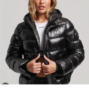 Svart glansig pufferjacka med huva - Säljer en svart, glansig pufferjacka med huva och dragkedja. Jackan är vadderad och har en oversized look, perfekt för kalla dagar. Den har långa ärmar och praktiska fickor framtill. Passar dig som vill ha en stilren och trendig vinterjacka.