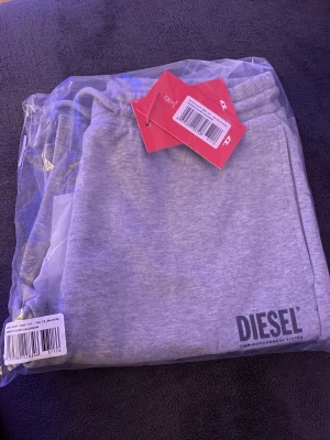 Grå mjukisshorts från Diesel - Snygga grå mjukisshorts från Diesel med logga på benet. Shortsen har elastisk midja med snörning och är tillverkade i mjuk bomullsmix för maximal komfort. Perfekta för en avslappnad och sportig stil.