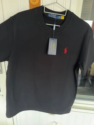 Svart t-shirt från Polo Ralph Lauren M - Svart t-shirt från Polo Ralph Lauren med klassisk rund hals och röd broderad logga på bröstet. Storlek L men passar bättre på M. Helt ny