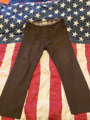 Bruna raka byxor från Hampton Republic - Snygga bruna byxor från Hampton Republic med klassisk rak passform. Byxorna har fem fickor, bälteshällor och knappstängning. Insidan är fodrad med rutigt tyg för extra stilpoäng. Perfekta för dig som gillar en clean och tidlös look. Det finns tecken på användning på byxorna men fortfarande använda i bra skick. Byxorna är i storlek 34/32. 