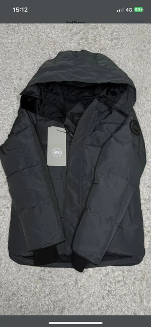 Svart pufferjacka från Canada Goose - Svart pufferjacka från Canada Goose med huva och klassisk logotyp på ärmen. Jackan har quiltad design, dragkedja framtill och ribbade muddar vid ärmslut. Materialet är polyester och dun, perfekt för kalla dagar. Snygg och stilren look som funkar till allt.