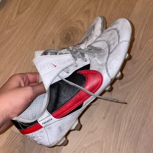 Nike Premier vita fotbollsskor - Nike Premier fotbollsskor i vitt med grå snörning och en stor röd swoosh på sidan. Skorna har svart detalj vid hälen och är tillverkade i skinn med perforerade partier för extra ventilation. Utrustade med dobbar för spel på gräs. Snygg och klassisk design för dig som gillar fotboll.