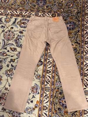 Beige Levi's 501 jeans W32 L32 - Säljer ett par klassiska Levi's 501 jeans i beige färg. Modellen har rak passform, fem fickor och knappgylf. Jeansen är tillverkade i 100% bomull och har den ikoniska läderpatchen bak i midjan. Perfekt för dig som gillar tidlös stil.