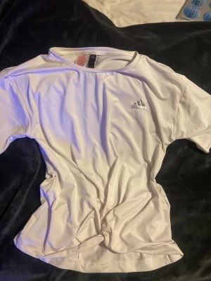 Vit Adidas t-shirt i polyester - Fräsch vit t-shirt från Adidas med klassisk logga på bröstet. Tillverkad i mjuk och lätt polyester som andas, perfekt för sport eller chill. T-shirten har rund hals och korta ärmar, stilren och enkel design som funkar till det mesta.