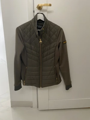 Olivgrön quiltad jacka från Barbour - Snygg olivgrön quiltad jacka från Barbour med guldfärgade dragkedjor och hög krage. Jackan har långärmade partier i mjukt material och quiltade paneler framtill och bak. Perfekt för dig som gillar stilren och sportig look.