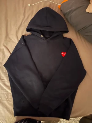 Navy blue Comme des Garçons hoodie med hjärta - Säljer en svart hoodie från Comme des Garçons Play med ikoniskt rött hjärta med ögon broderat på bröstet. Tröjan har huva, långärmad design och är tillverkad i mjuk bomull. Perfekt för dig som gillar streetwear och vill ha något stilrent med en twist.