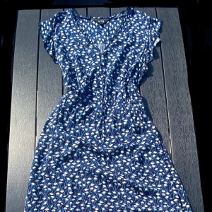 Blå långklänning med blommigt mönster - Säljer en blå långklänning med vitt och grönt blommigt mönster. Klänningen har korta ärmar, v-ringning och ett knytband i midjan. Klänningen är extra lång och är anpassad för långa personer. Perfekt för varma dagar och har en luftig passform. Materialet känns mjukt och bekvämt.