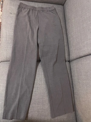 Grå kostymbyxor från Åhléns, stl 38 - Snygga grå kostymbyxor från Åhléns i rak modell med resår i midjan för extra komfort. Byxorna är tillverkade i ett mjukt material som ger en clean och stilren look. Perfekta att matcha med en enkel topp eller skjorta.