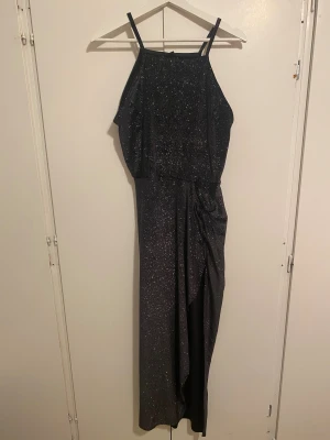 Glittrig svart långklänning med slits - Supersnygg svart långklänning med tunna axelband och glittrigt tyg. Klänningen har en cool drapering i midjan och en hög slits framtill som ger extra edge. Perfekt för dig som vill sticka ut med en glamorös look. Materialet känns mjukt och följsamt.
