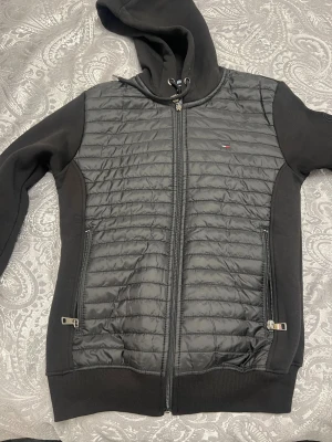 Svart pufferjacka Tommy Hilfiger S - Svart pufferjacka från Tommy Hilfiger i storlek S. Jackan har quiltad framsida i syntetmaterial, mjuka ärmar och huva med snörning. Två sidofickor med dragkedja och diskret logga på bröstet och ärmen. Perfekt för kyliga dagar.
